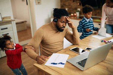 Small black girl pulling father's arm while he is working on a computer at home. Mother and son are doing homework in the background. 