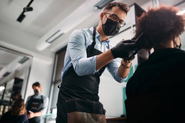 Hairdresser with protective face mask brushing black woman's hair during appointment at the salon.