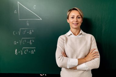 Confident teacher standing with arms crossed in front of blackboard and looking at camera. 