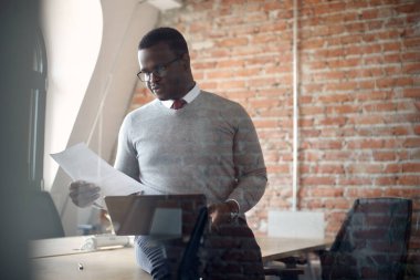 Black male entrepreneur using laptop while reading business reports in the office. The view is through the glass. 