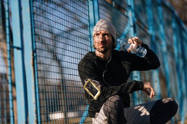 Young sportsman taking a break and drinking water while having sports training outdoors.