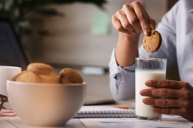 Close-up of freelance worker having a break and dipping cookie in a glass of milk. 