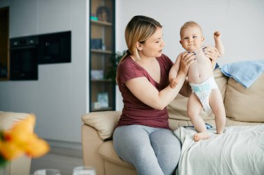 Young mother holding her small son who is trying to stand on the sofa at home.