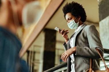 African American businesswoman wearing face mask while using voice assistant on mobile phone at public train station. 