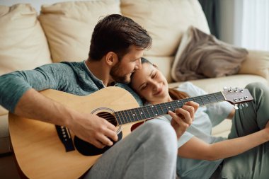 Young man playing acoustic guitar while wife is leaning on his shoulder at home.