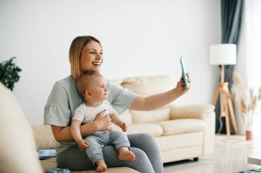 Happy mother using smart phone while taking selfie with her baby son at home.