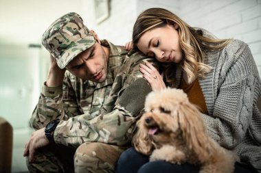 Young distraught soldier and his wife feeling worried about his deployment for military assignment while sitting with a dog at home. 