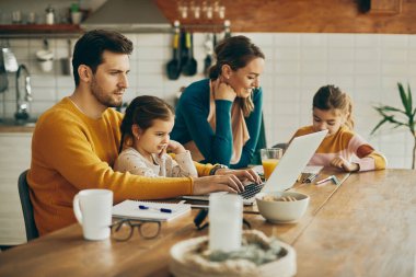 Working father using computer while daughter is sitting on his laptop at home. Mother and daughter are doing homework in the background. 