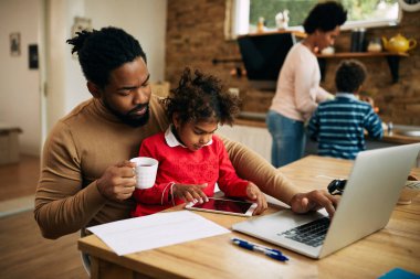 Black working father using laptop while daughter is sitting on his lap and using touchpad at home. 