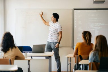 Computer science teacher talking in front of whiteboard to group of high school students in the classroom. 