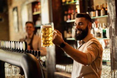 Young happy bartender holding a glass of draft beer while working in a pub. 