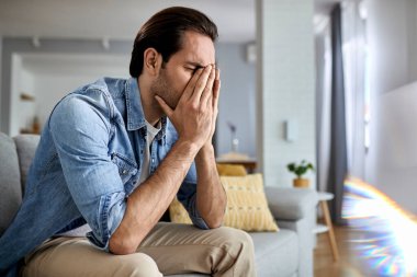 Young man feeling stressed and having headache while sitting alone in the living room. 