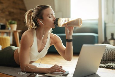Genç sporcu kadın evde dizüstü bilgisayar kullanırken yerde dinleniyor ve smoothie içiyor.. 