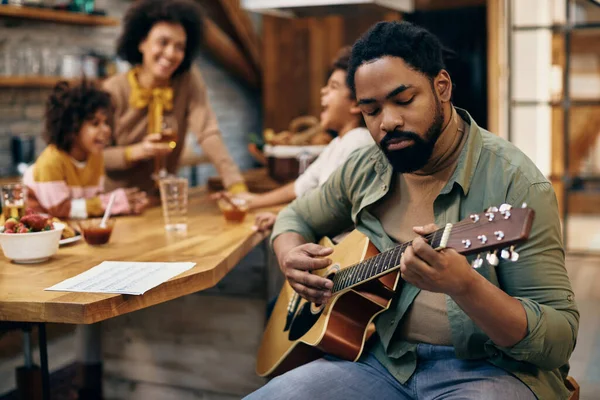 Black man playing acoustic guitar while spending time with his family at home.