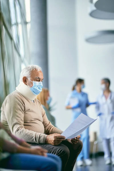 Senior man with neck injury wearing cervical collar and reading his medical reports in waiting room at the hospital. He is wearing face mask due to COVID-19 pandemic.