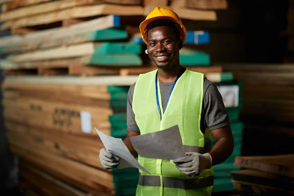 Happy African American worker going through delivery plans at lumber distribution warehouse and looking at camera.