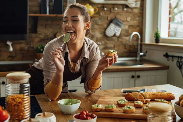 Young woman tasting food with eyes closed while preparing avocado bruschetta in the kitchen.