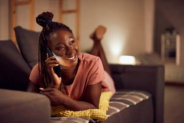 Young happy black woman using smart phone and making a phone call while relaxing on the sofa at night at home.
