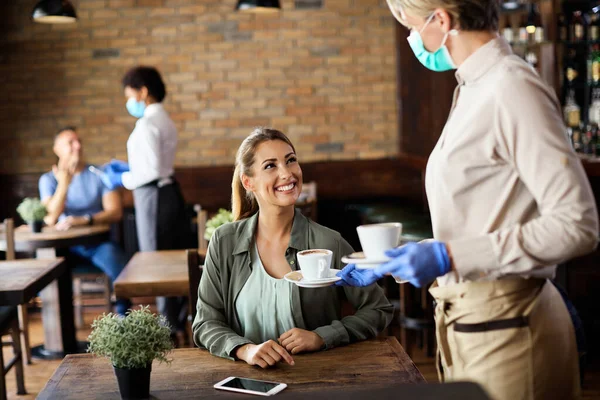 Waitress with protective face mask and gloves serving coffee to happy woman in a cafe. Focus is on happy woman.