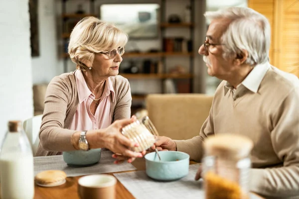 Senior couple communicating while eating breakfast in the morning. Focus is on woman pouring food in husband\'s bowl.