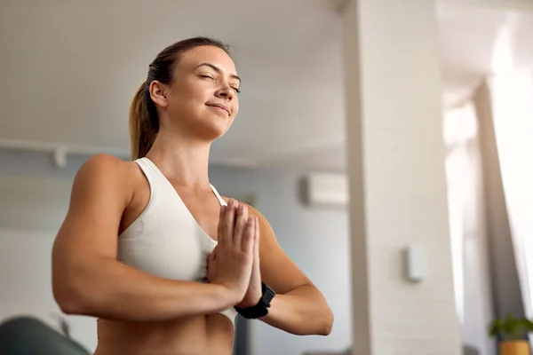 Low angle view of athletic woman with hands clasped doing breathing exercise at home.