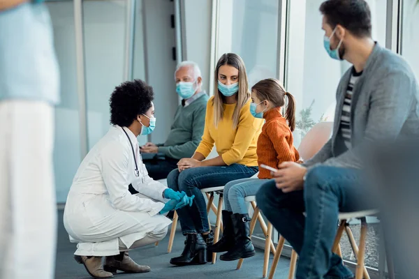 Black female pediatrician communicating with little girl and her mother in a waiting room at medical clinic. All of them are wearing face masks due to coronavirus pandemic. 