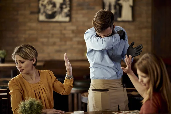 Young waiter coughing into elbow while serving customers in a pub.