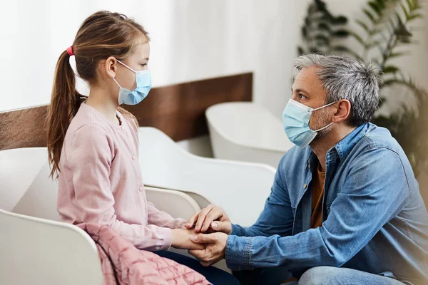 Caring father encouraging his daughter while waiting for dental appointment at dentist\'s office. They are wearing face masks due to coronavirus pandemic.