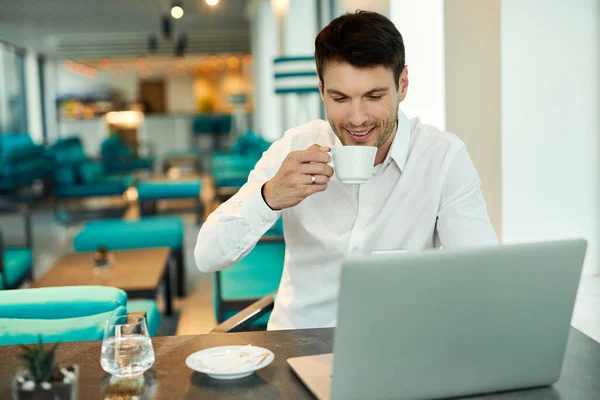 Smiling entrepreneur surfing the net on laptop while drinking coffee in a cafe.