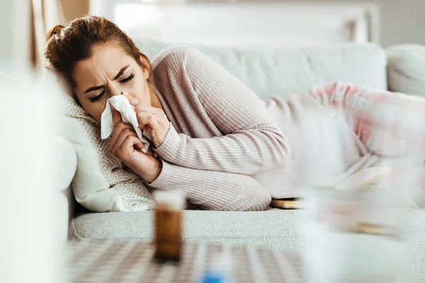 Young woman with flu virus lying down on sofa and blowing her nose.