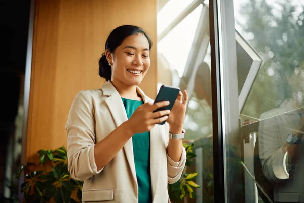 Happy Asian Businesswoman Text Messaging Cell Phone While Standing Window — Zdjęcie stockowe