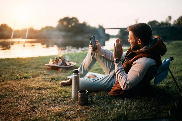 Happy man using mobile phone and waving while talking to his wife and camping in nature. Copy space.