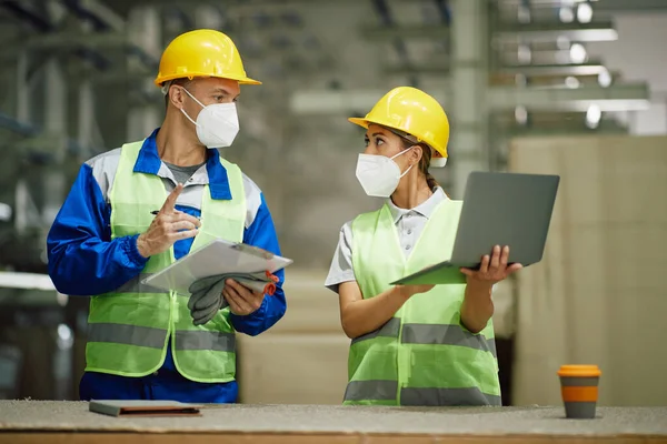 Warehouse workers using laptop and analyzing delivery schedule while wearing face masks in a storage room.