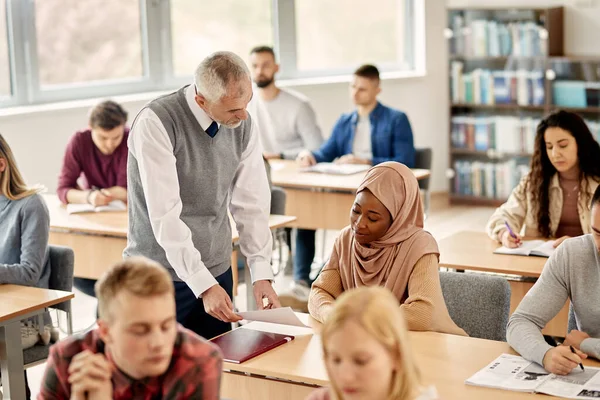 Mature teacher communicating with black Islamic student while assisting her at university classroom.