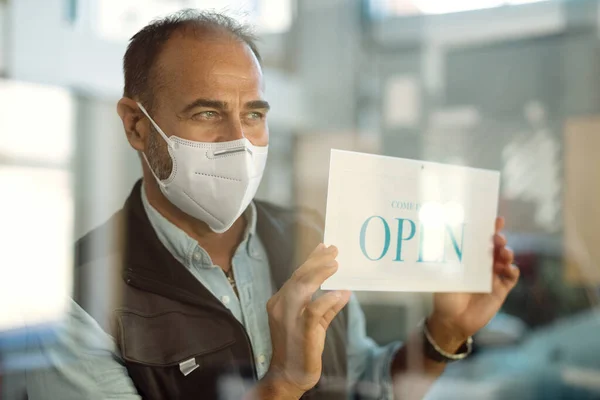 Auto mechanic wearing face mask while hanging open sign on entrance door. The view is through the glass.
