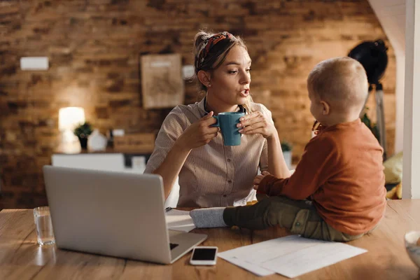 Single mother drinking coffee and communicating with her small son while working at home.
