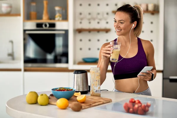 Happy athletic woman drinking smoothie while using mobile phone and listening music over ear phone in the kitchen.