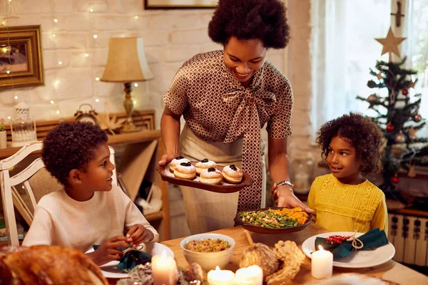 Happy African American mother bringing food at dining table during Christmas meal with her kids at home.