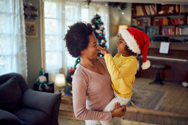 Happy African American mother holding her small daughter who is touching her nose on Christmas day at home.