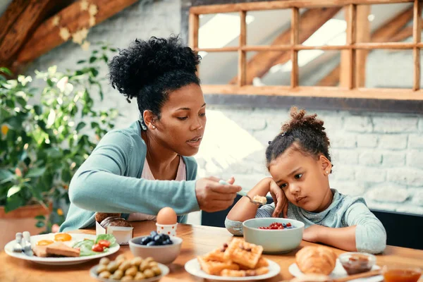 African American mother feeding her small daughter who is refusing to eat her breakfast at dining table.