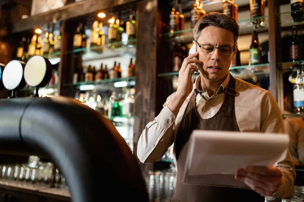 Bartender reading check list while talking on mobile phone and working in a pub.