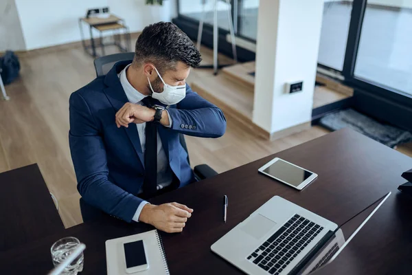 Businessman with protective face mask sneezing into elbow while working in the office.