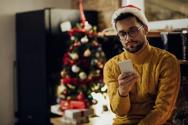 Young sad man texting on smart phone while feeling lonely on Christmas Eve at home.