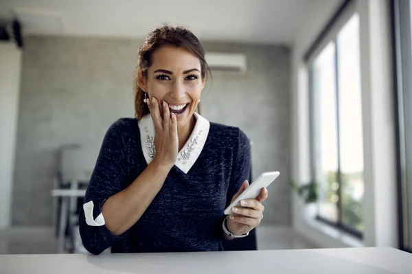 Excited businesswoman online dating over mobile phone and looking at camera in the office.