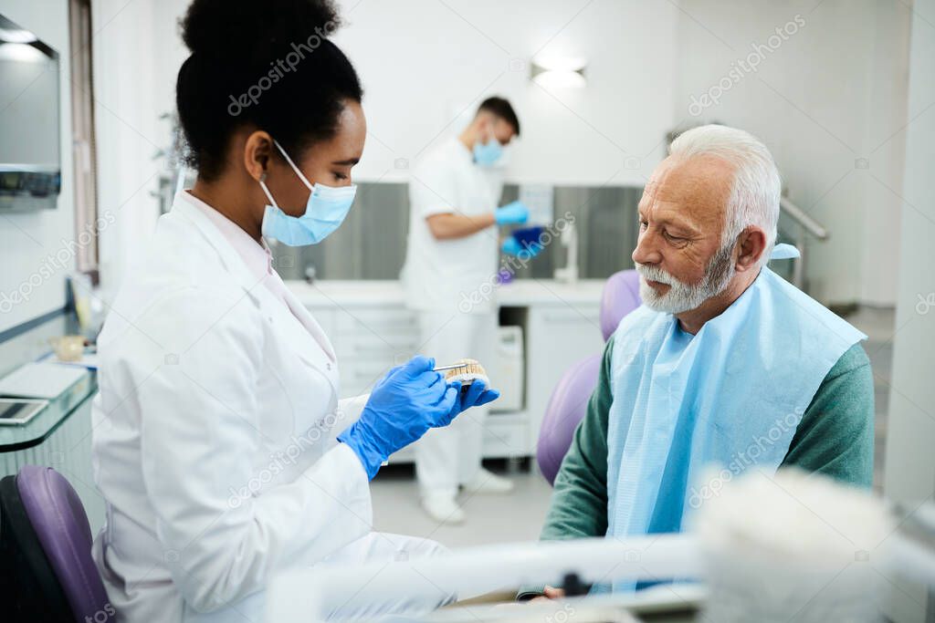 African American dentist using dental model while talking to mature patient during dental procedure at dentist's office.