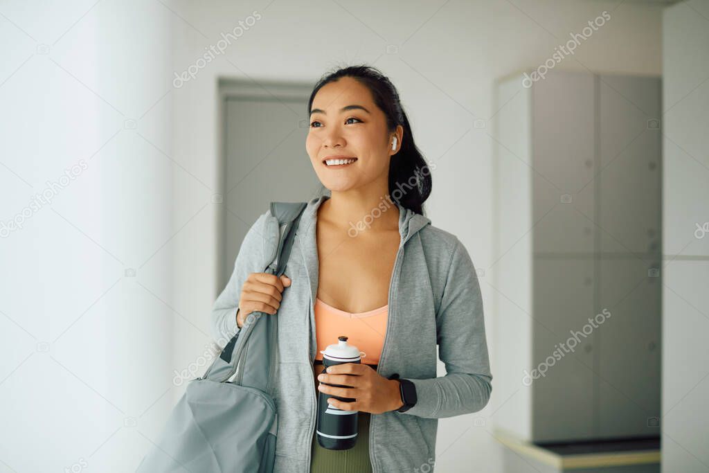 Young happy Asian sportswoman standing in locker room at the gym and looking away.