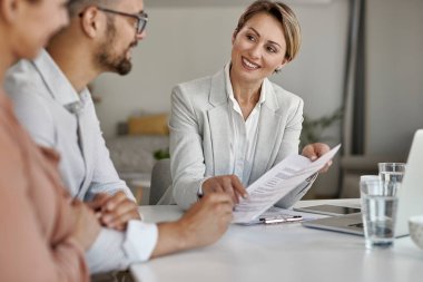 Happy insurance agent talking to a couple and offering them to sign a contract during a meeting. 