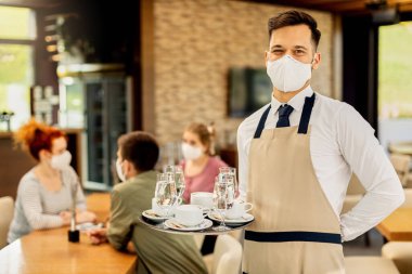 Happy waiter serving coffee to his customers while wearing protective face mask in a cafe due to coronavirus epidemic. 