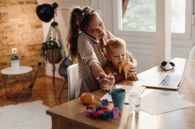 Stay at home mother talking on the phone while working at feeding her son with a banana. 