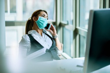 Female entrepreneur with face mask communicating over cell phone while working in the office. 